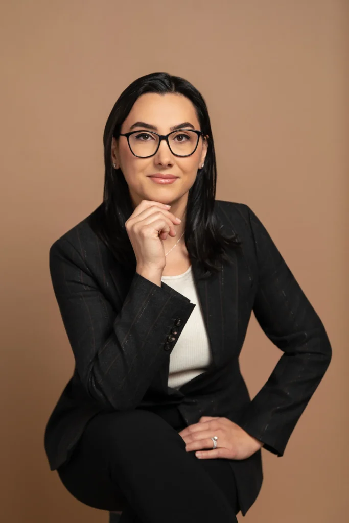 Karine Akopchikyan, attorney, seated in a studio portrait wearing glasses and a dark blazer, resting her chin on her hand against a neutral tan background.